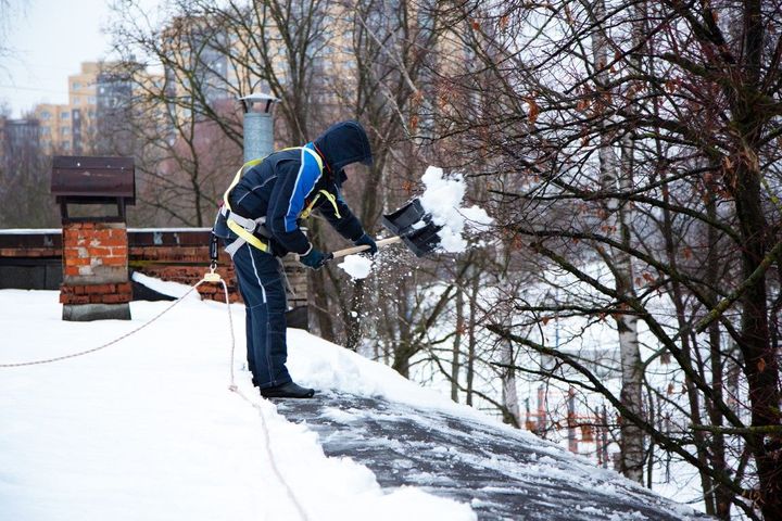 Worker shoveling snow from a roof near bare trees and buildings on a winter day