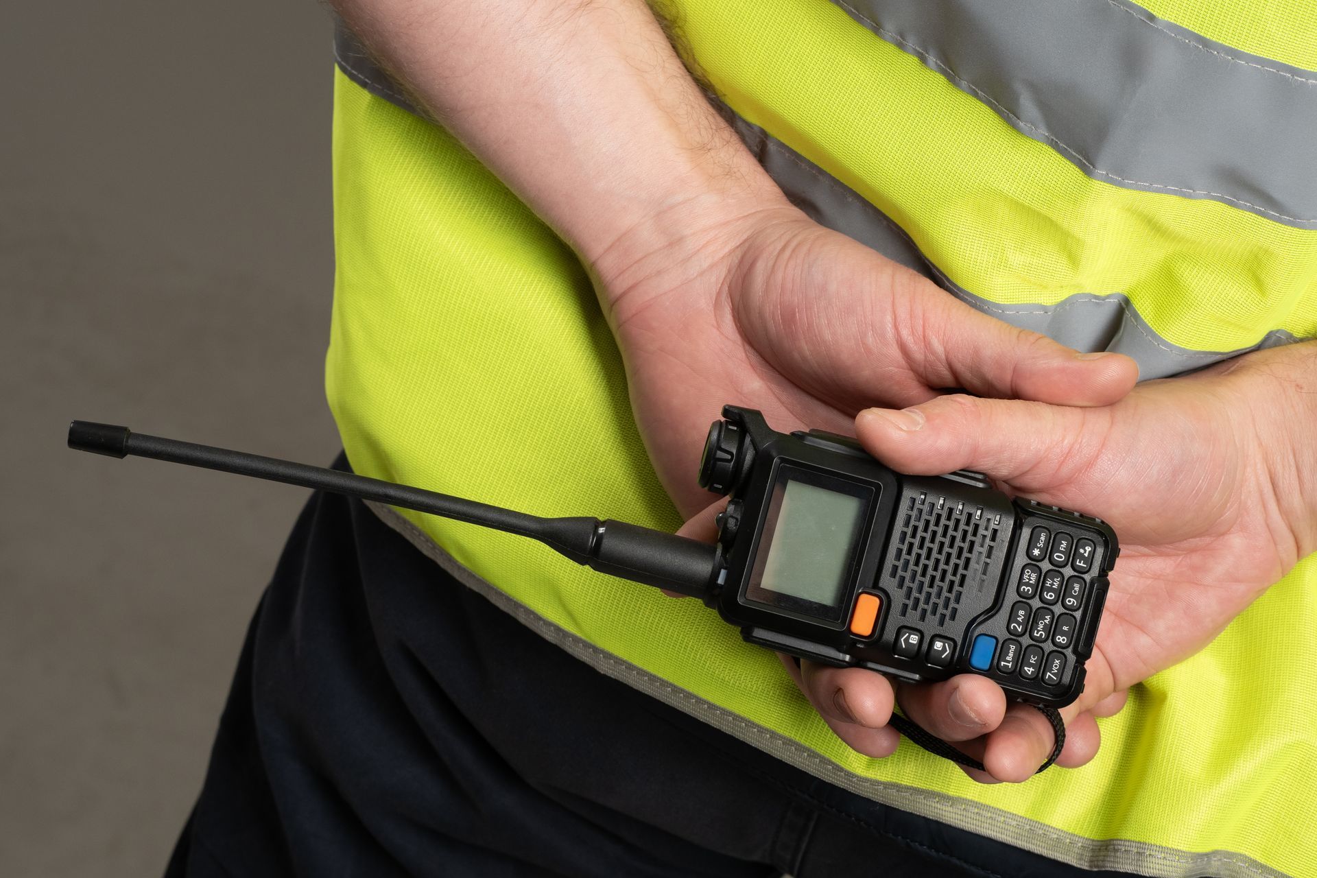 Person holding a black walkie-talkie with long antenna and orange button against a yellow vest