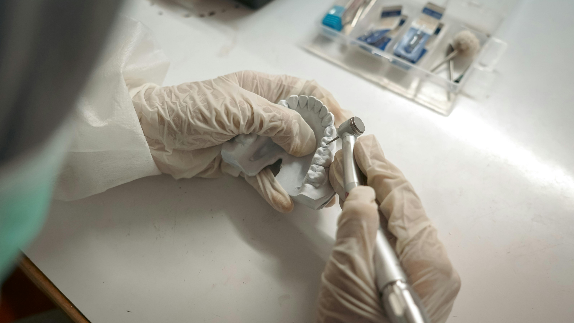 Gloved hands handling a dental tool over a white work surface with small medical items nearby