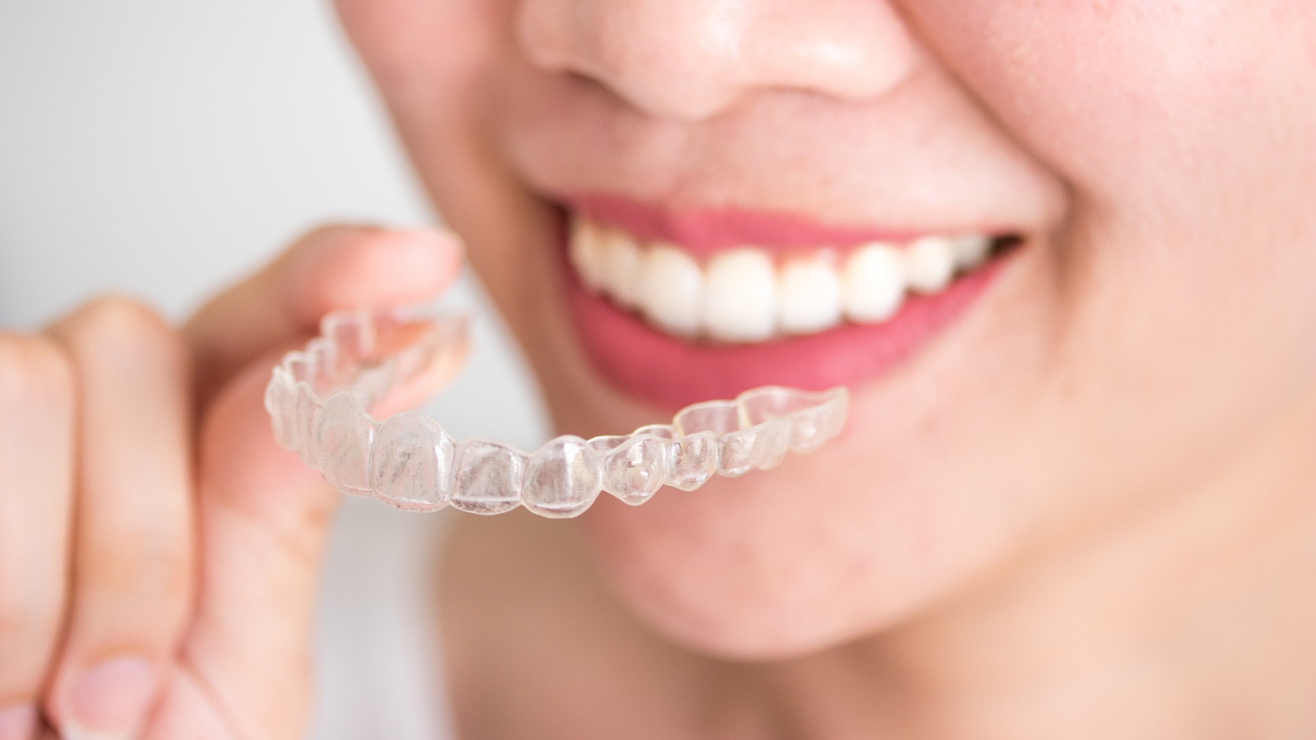 Person smiling while holding a clear orthodontic aligner close to their teeth