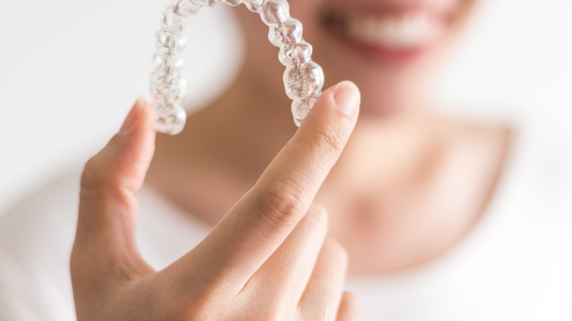 Close-up of a hand holding a clear retainer in front of a smiling face