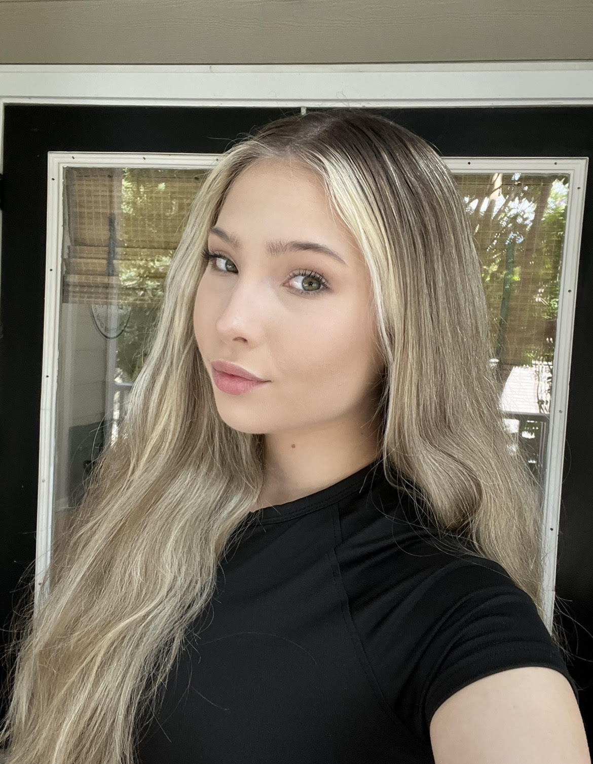 A person with long, two-toned blonde and brown hair in a center part, looking at the camera against a windowed door.