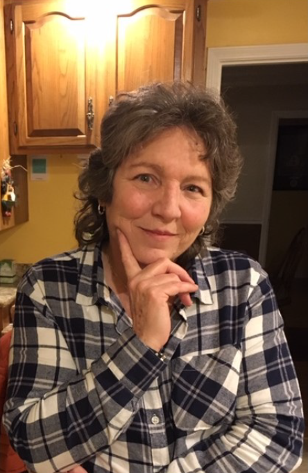 A person wearing a blue and white plaid shirt poses for a portrait with their hand to their chin in a kitchen.