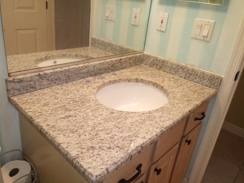 Bathroom vanity with a granite countertop and a white, round sink. Light wood cabinet, black hardware.