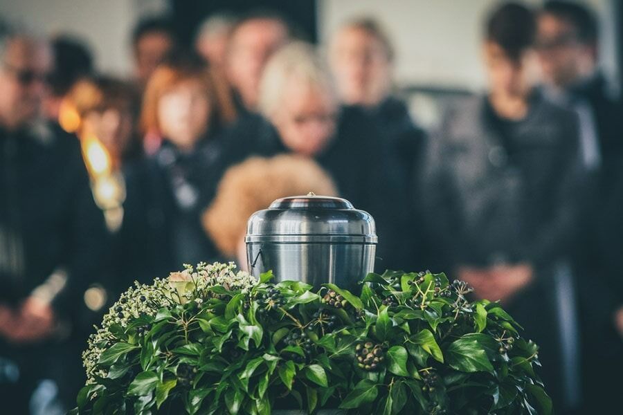 Urn Atop a Green Wreath With Mourners in the Background — Finlayson & McKenzie Funeral Directors in Rockhampton City, QLD