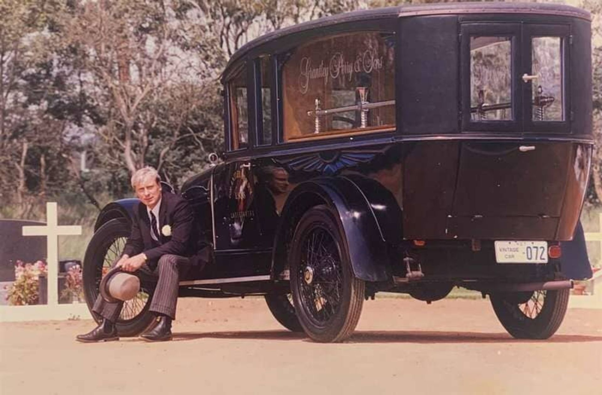 Man in a Suit Sits Beside a Vintage Black Hearse — Finlayson & McKenzie Funeral Directors in Rockhampton City, QLD