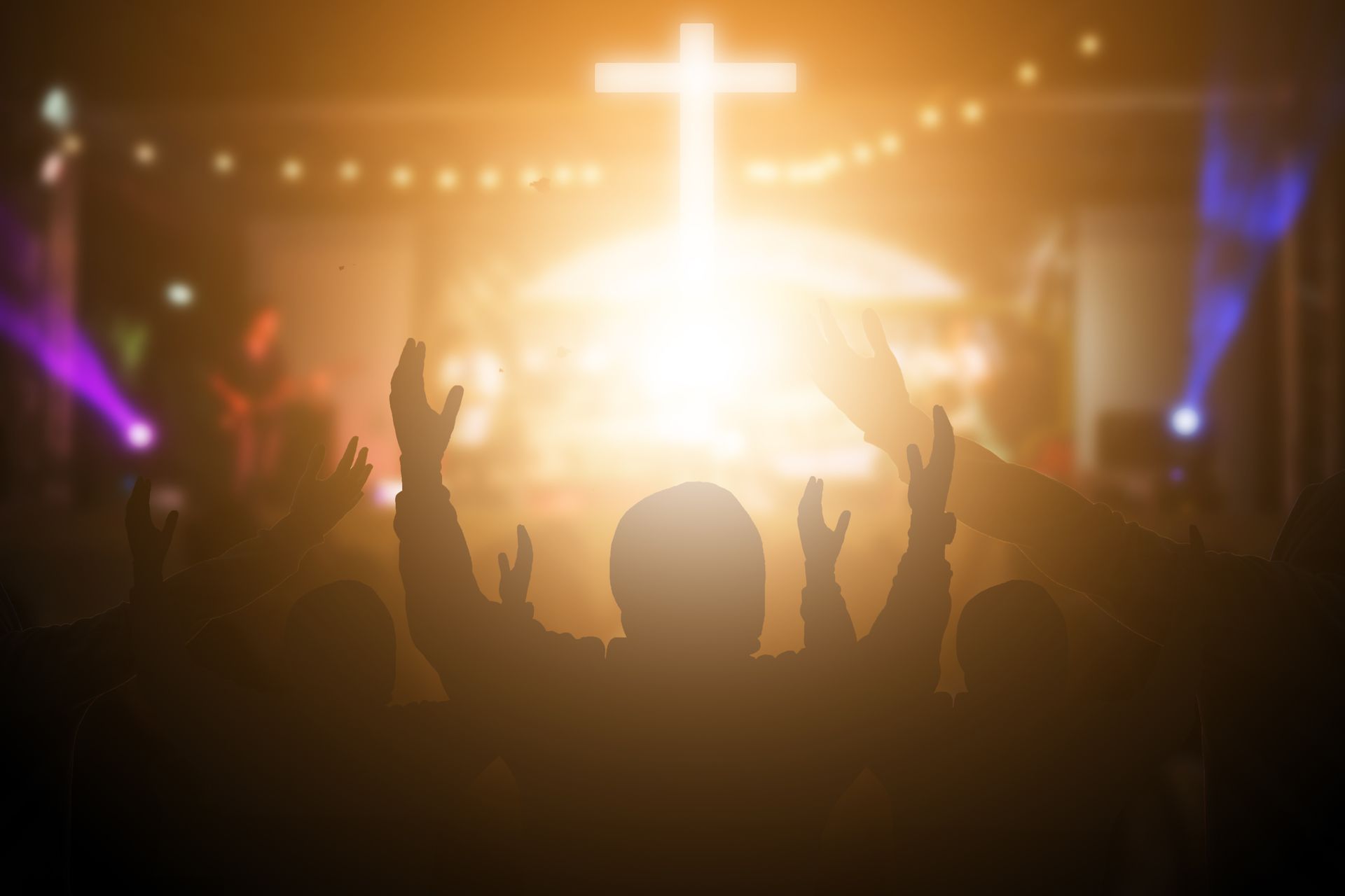 Crowd worshipping before a glowing cross on a stage with raised hands and colorful lights