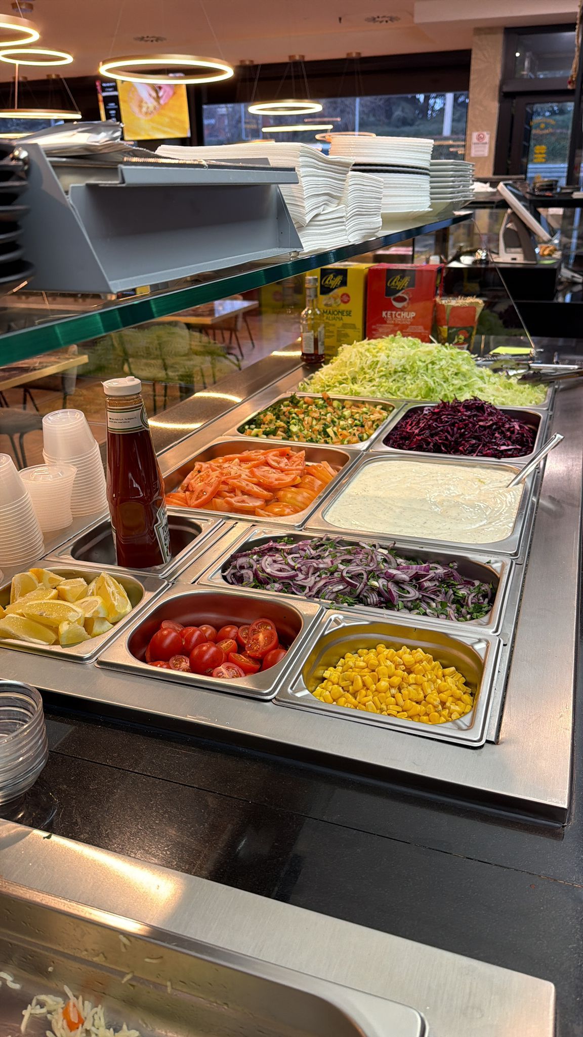 Buffet di insalate con vassoi di verdure tritate, lattuga e condimenti, protetto da una barriera di vetro parasputi.