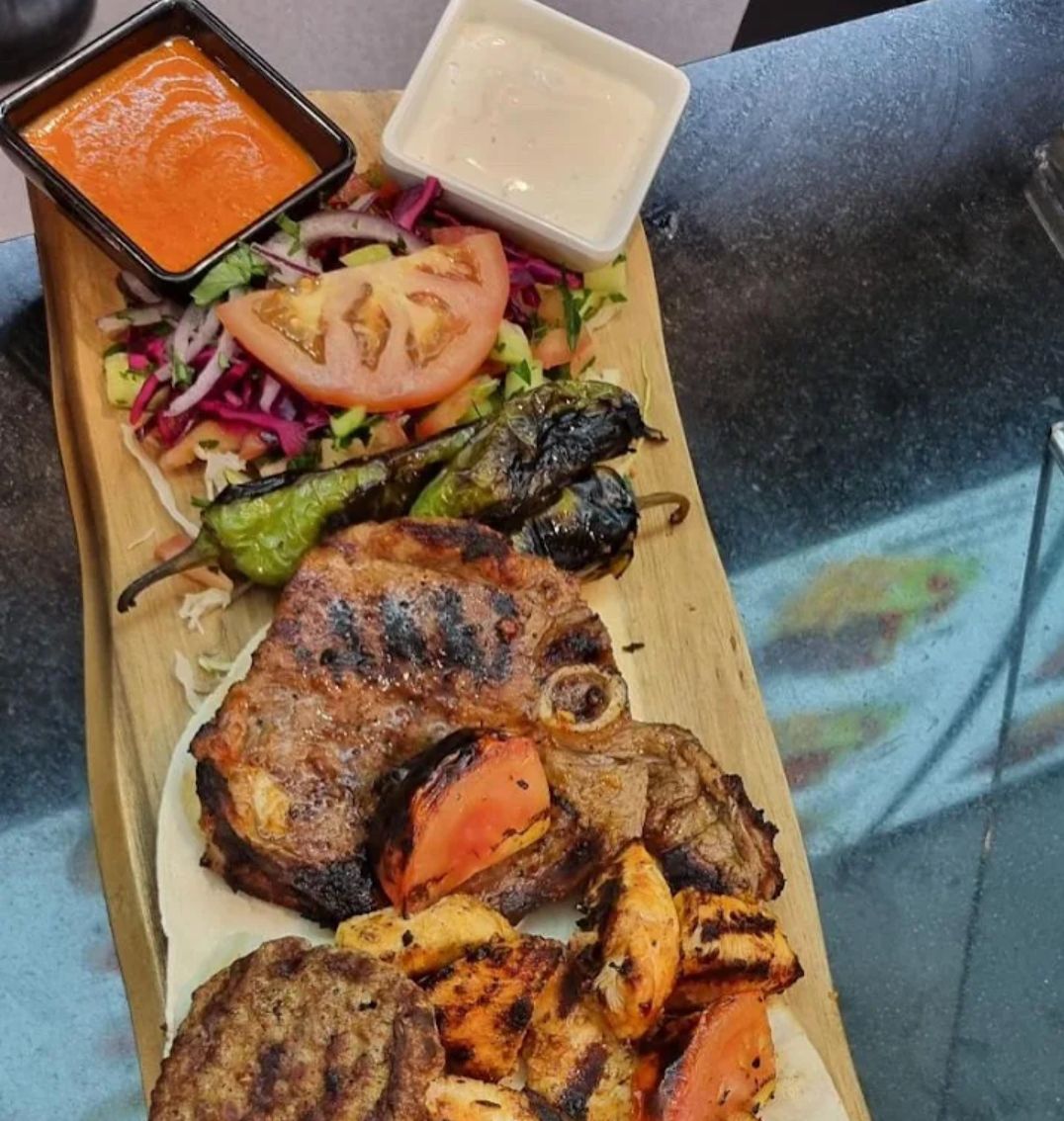 Piatto di carne alla griglia con insalata, verdure grigliate e due salse, servito su un tagliere di legno.