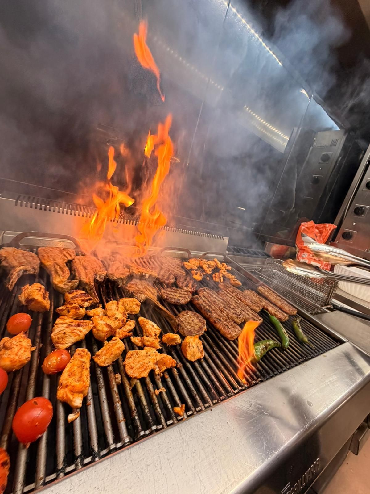Carne e verdure grigliate che sfrigolano su un barbecue a cielo aperto, con fiamme e fumo che si alzano