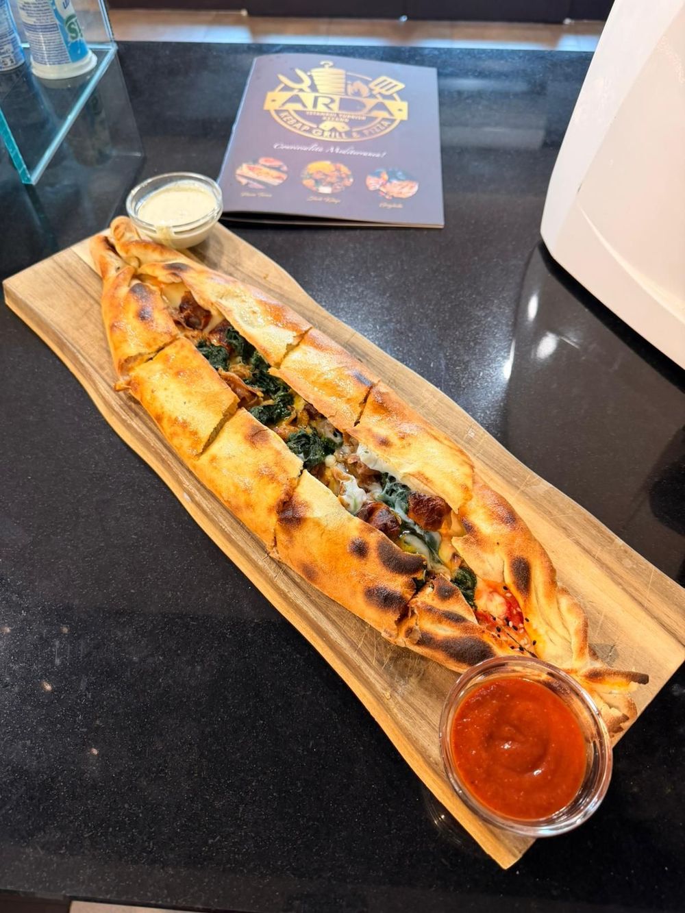 Un lungo sandwich grigliato su un vassoio di legno con salse per intingere su un tavolo nero.
