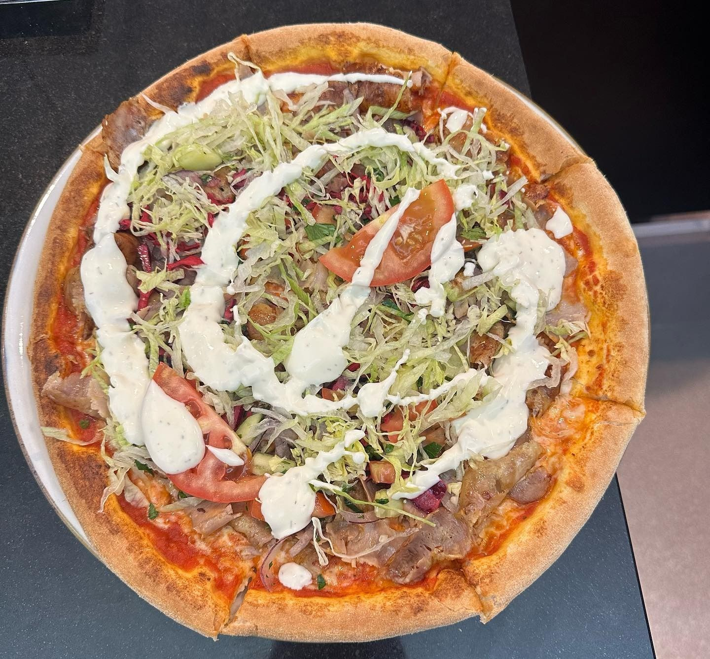 Una pizza rotonda condita con lattuga tritata, pomodoro, carne e un filo di salsa bianca, servita su un vassoio scuro.