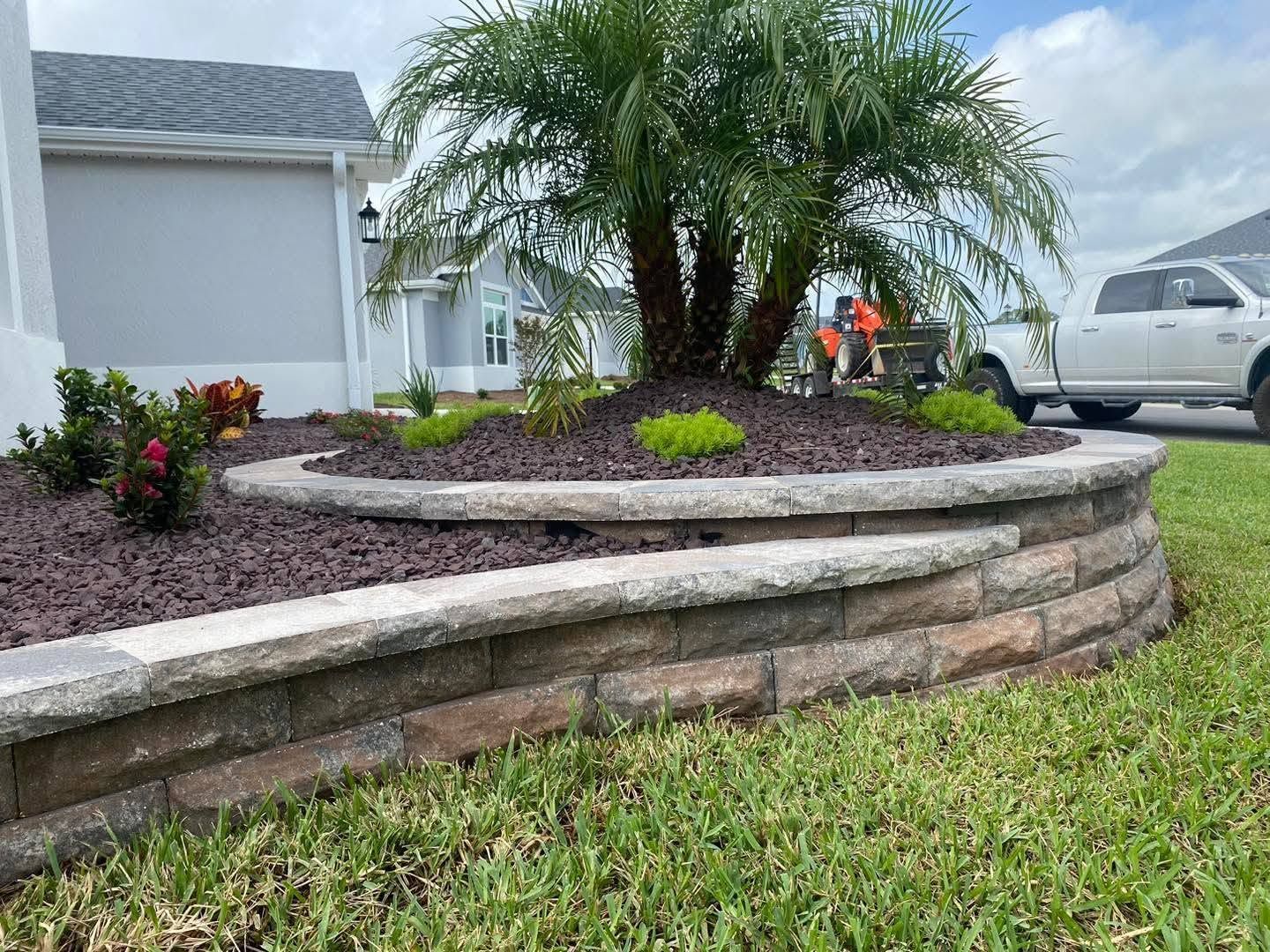 Landscaped front yard with palm tree, stone retaining wall, and a worker near a white pickup truck