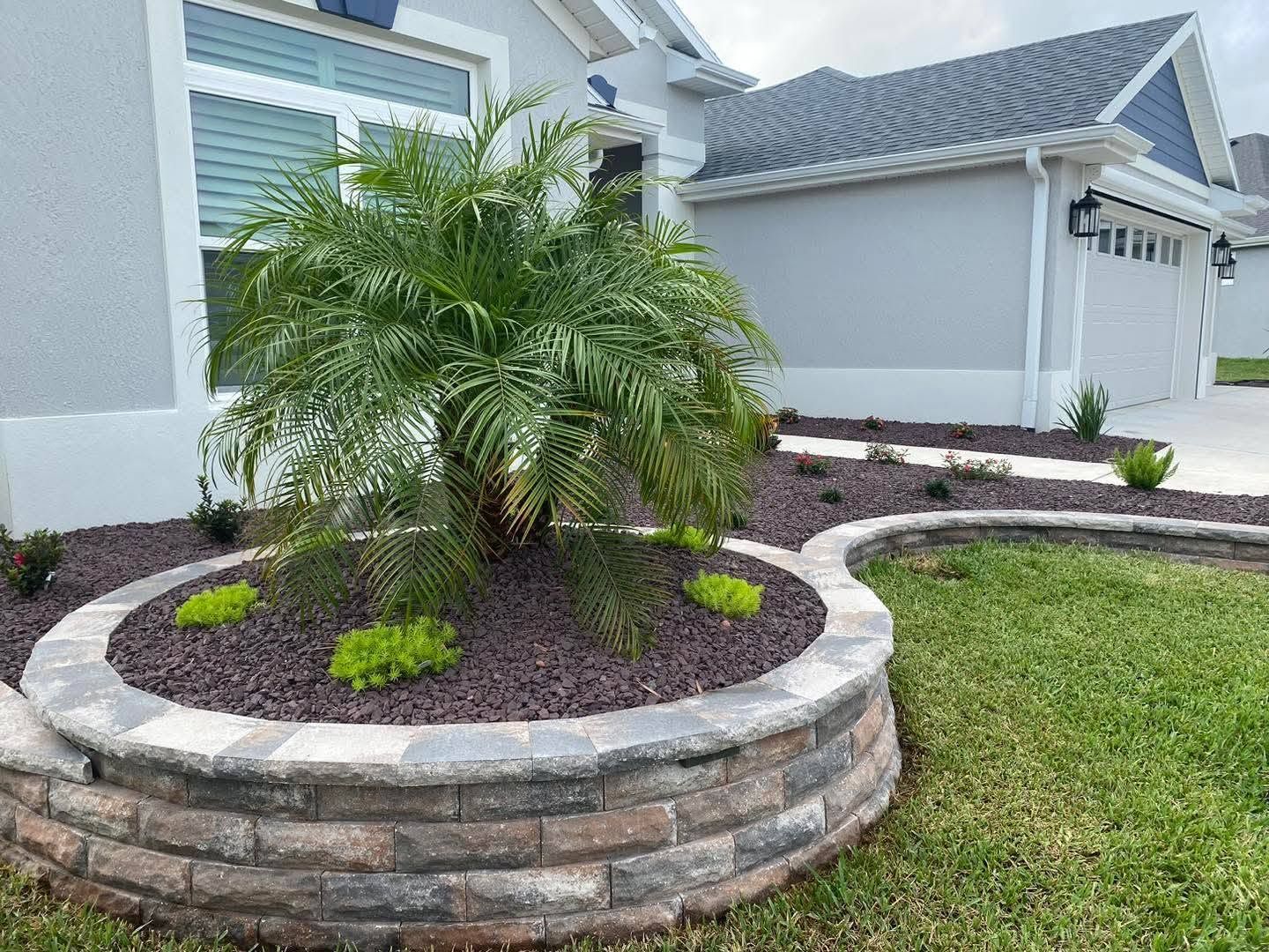 Raised stone planter with a palm tree and mulch in front of a suburban house