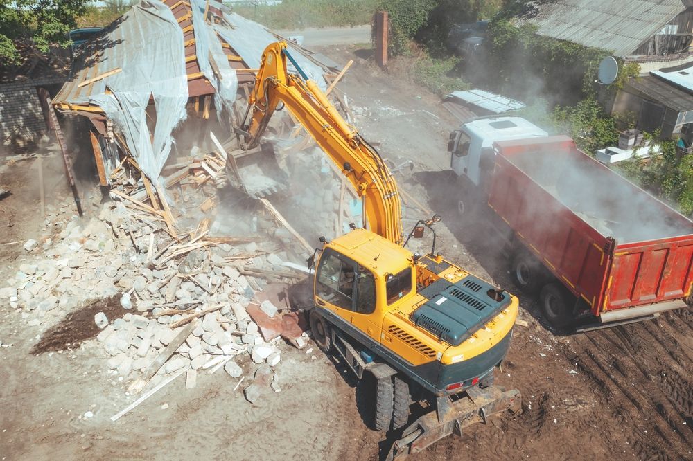 Excavator demolishing a collapsed building beside a red dump truck in a dusty worksite