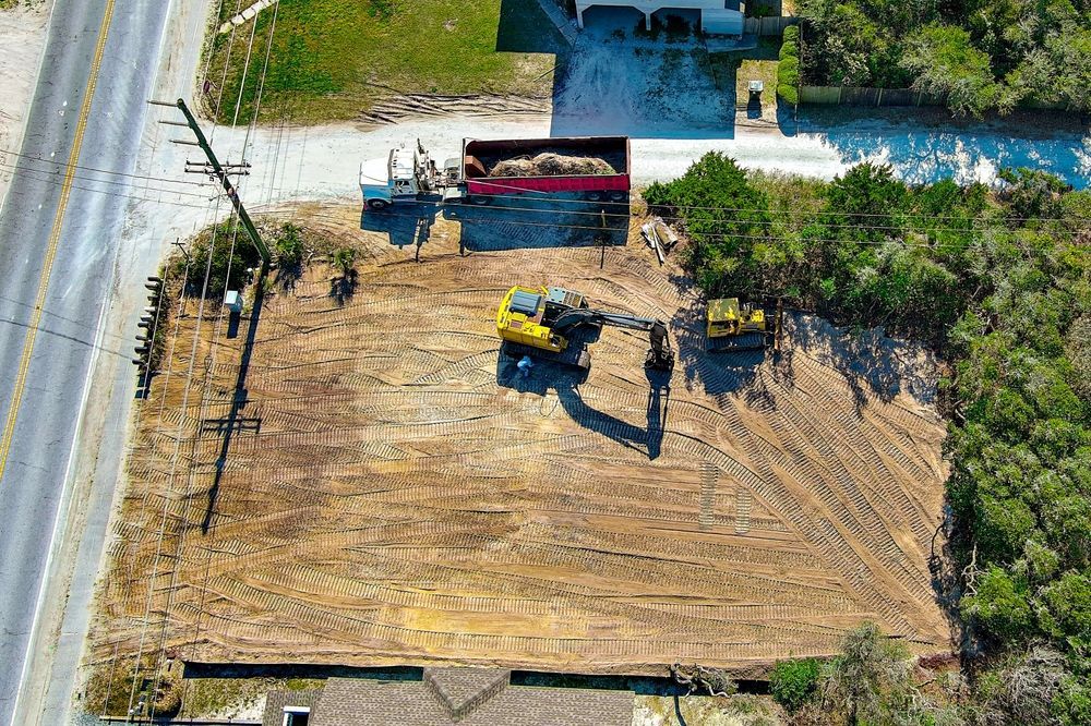 Aerial view of a construction site with a bulldozer on a dirt lot beside a road and trees.