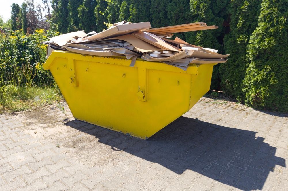 Yellow dumpster filled with cardboard scraps on a paved driveway beside green hedges