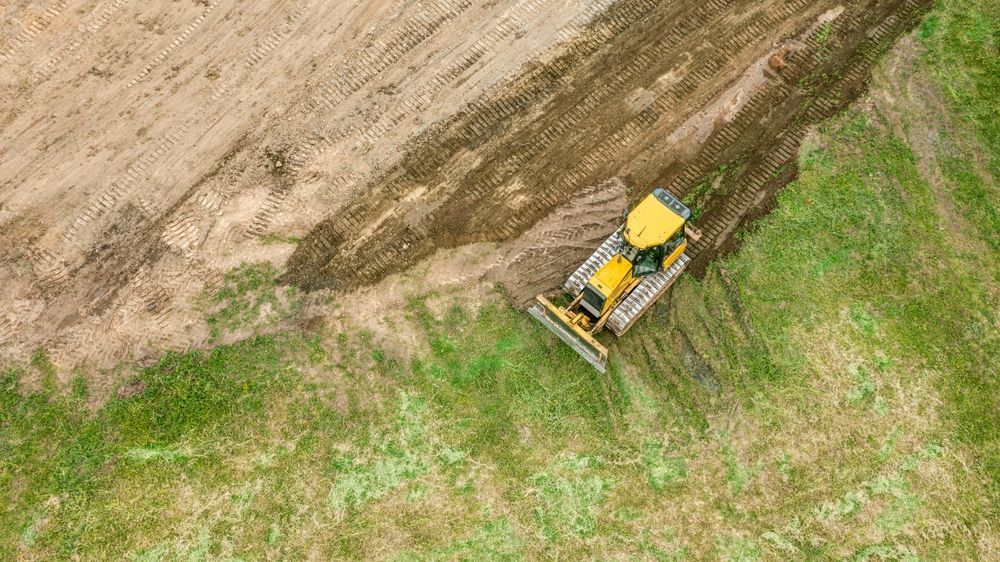 Yellow tractor plowing a muddy field at the edge of green grass, seen from above