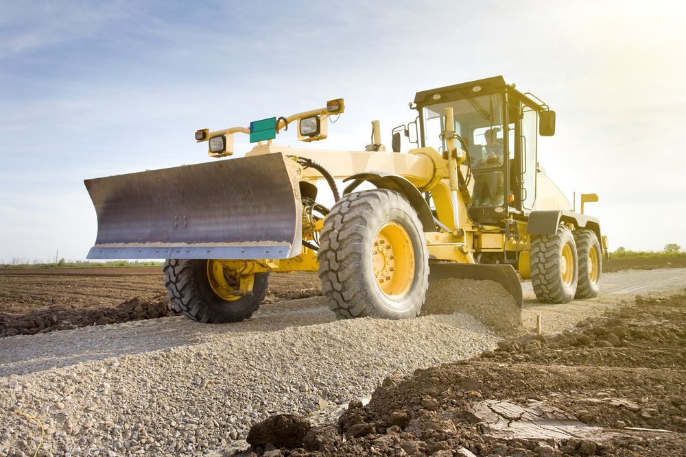 Yellow road grader leveling gravel on a dirt road at sunset