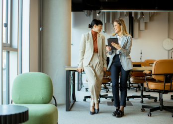 Two people walking and talking in a bright hallway, holding coffee cups