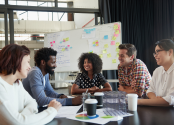 Team meeting in a bright office with five people seated around a table and a whiteboard behind them