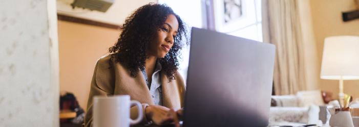 a woman with curly hair sits at a laptop in a living room, looking thoughtfully at the screen