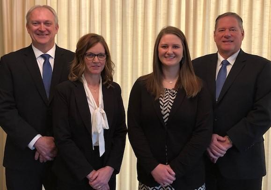 Four professionals stand side-by-side in formal business attire in front of a neutral curtain backdrop.
