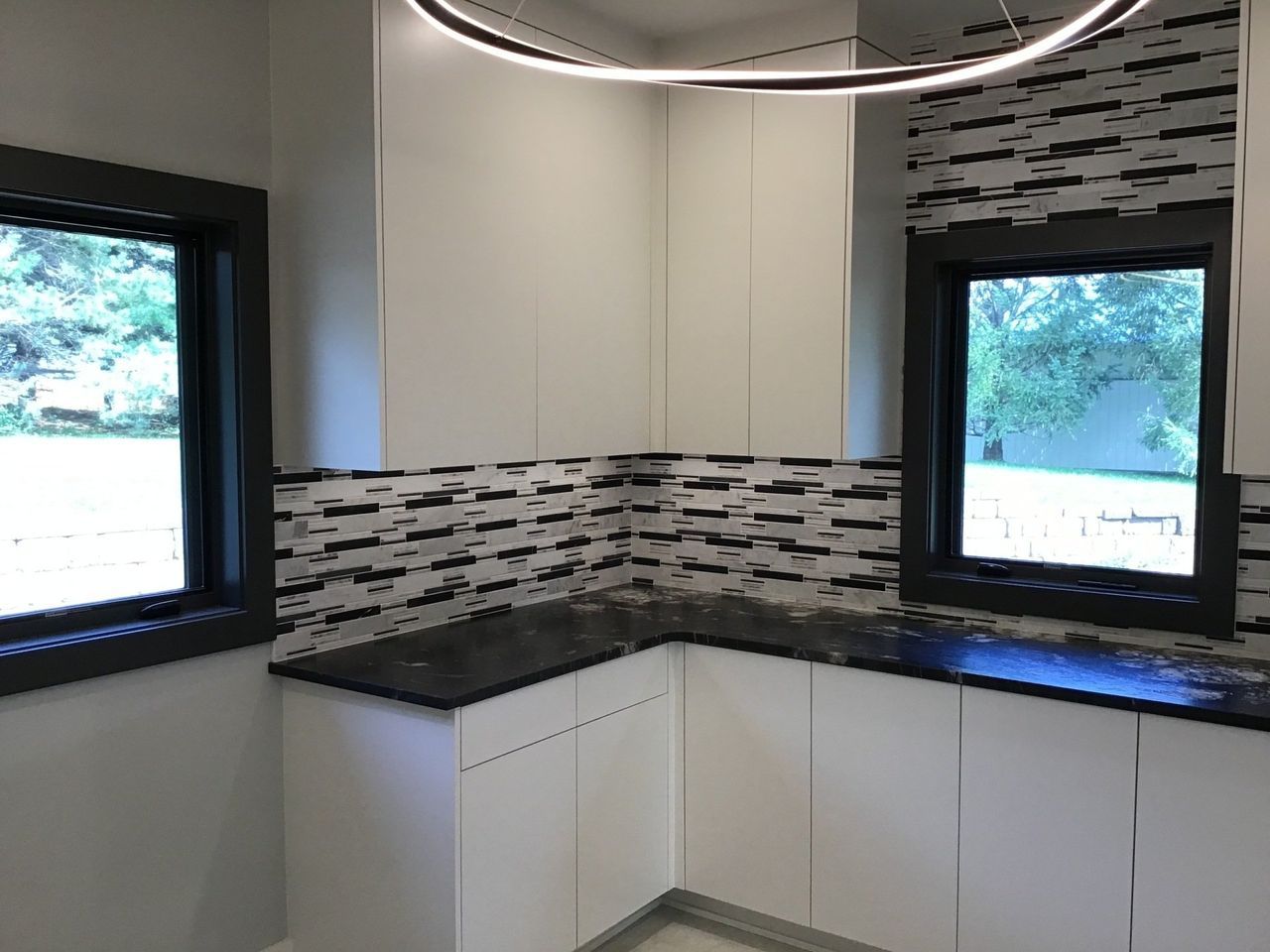 Modern kitchen corner with white cabinets, dark countertops, mosaic backsplash, and two windows