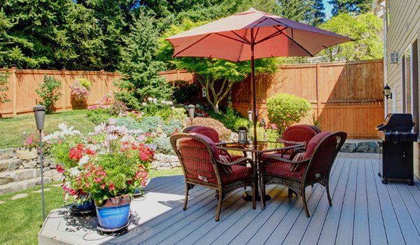 Backyard patio with wicker dining set, red umbrella, potted flowers, and grill on a gray deck
