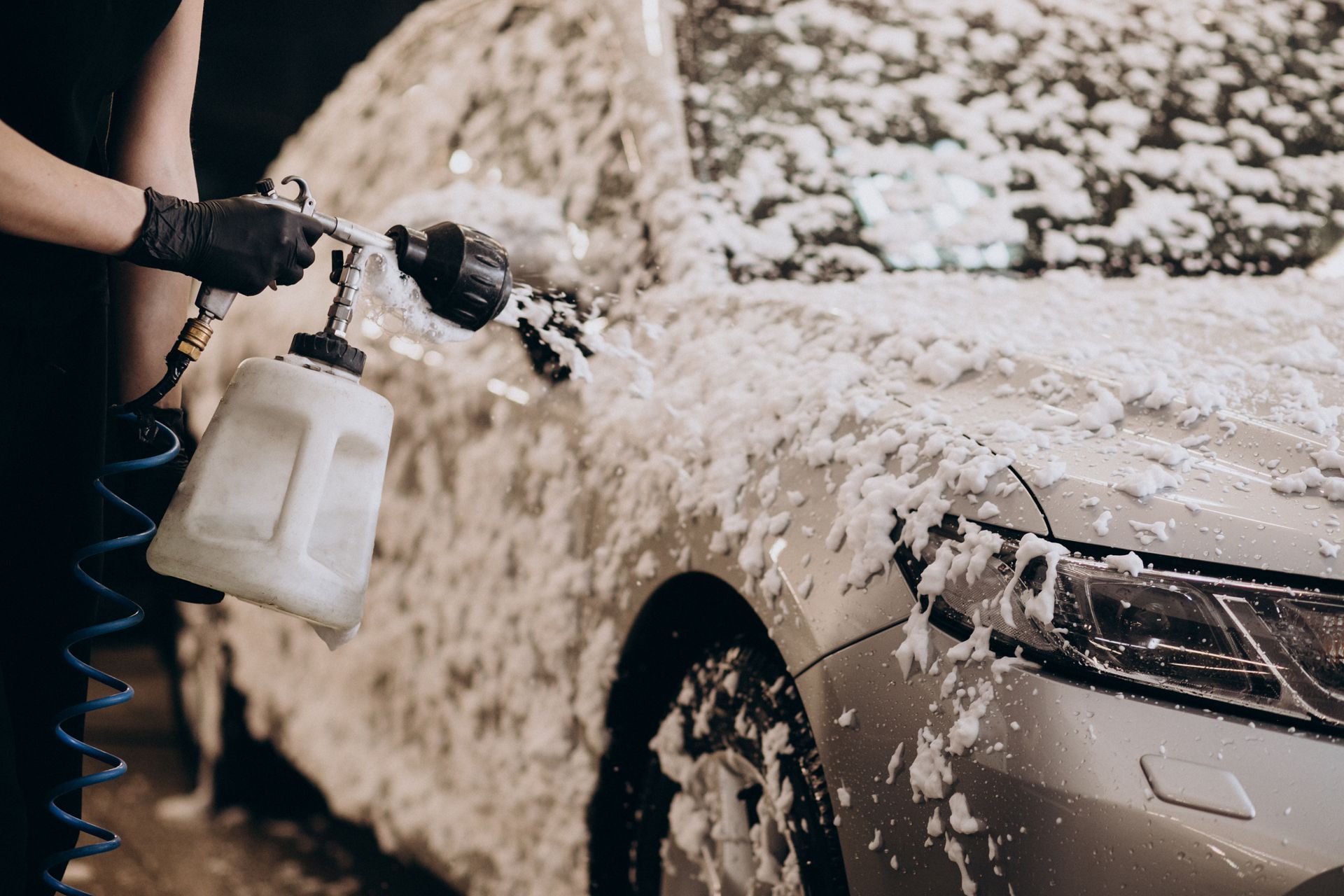 A gloved hand uses a spray gun to apply white cleaning foam onto the side of a silver car.
