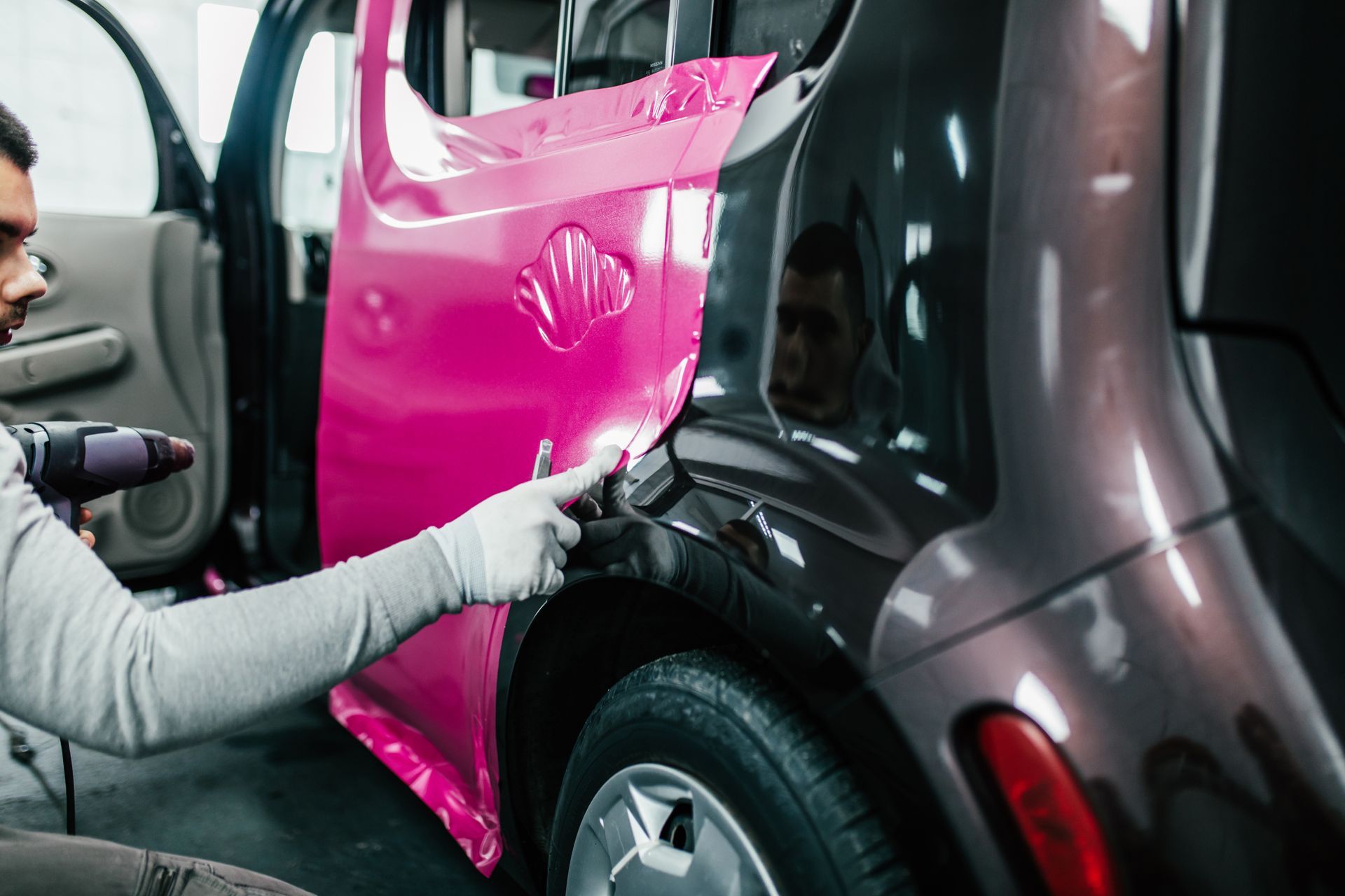 A person in gloves uses a heat gun to apply a bright pink vinyl wrap to the side of a car in a shop.