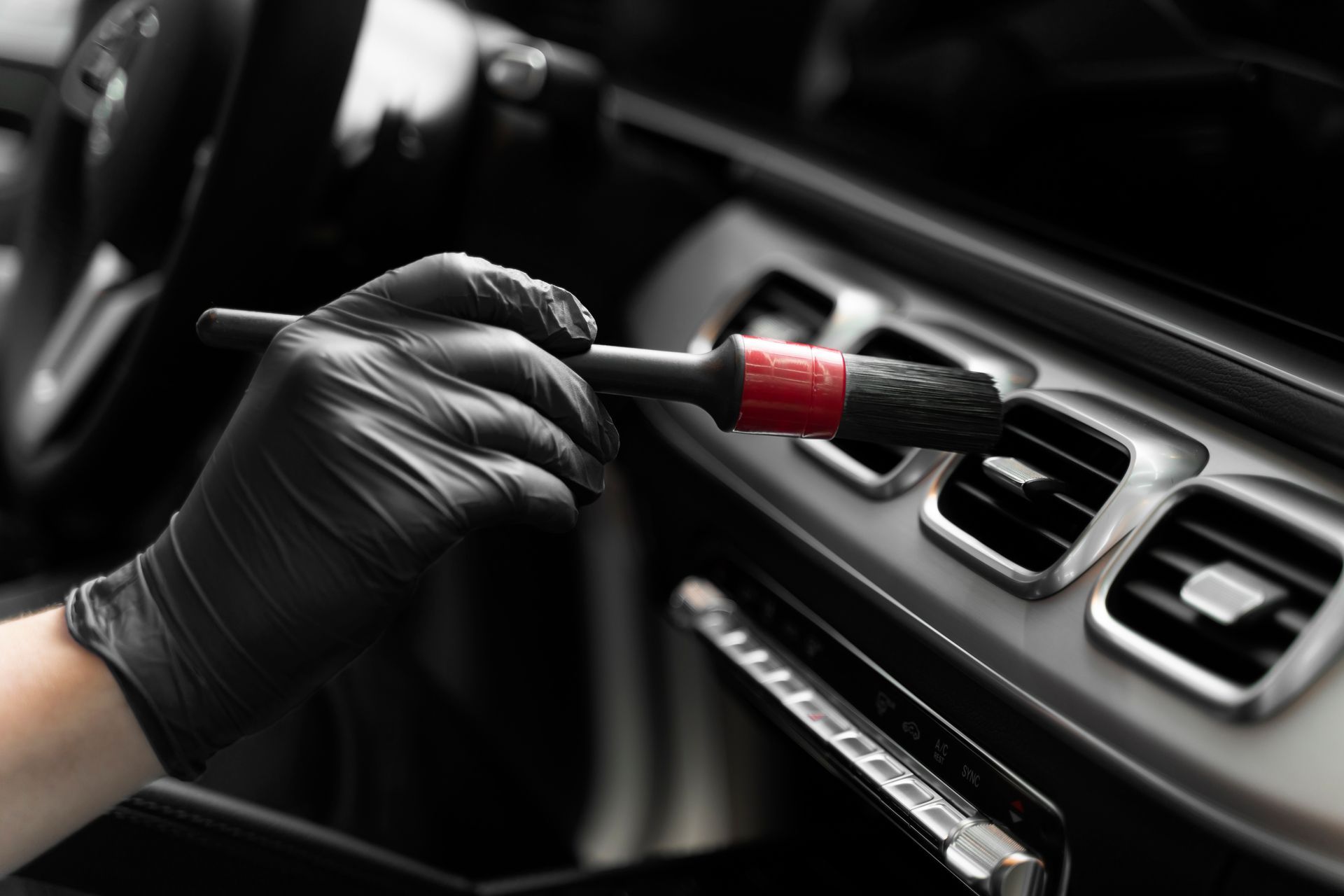 A hand wearing a black glove uses a small red-handled brush to clean the air vent of a car interior.
