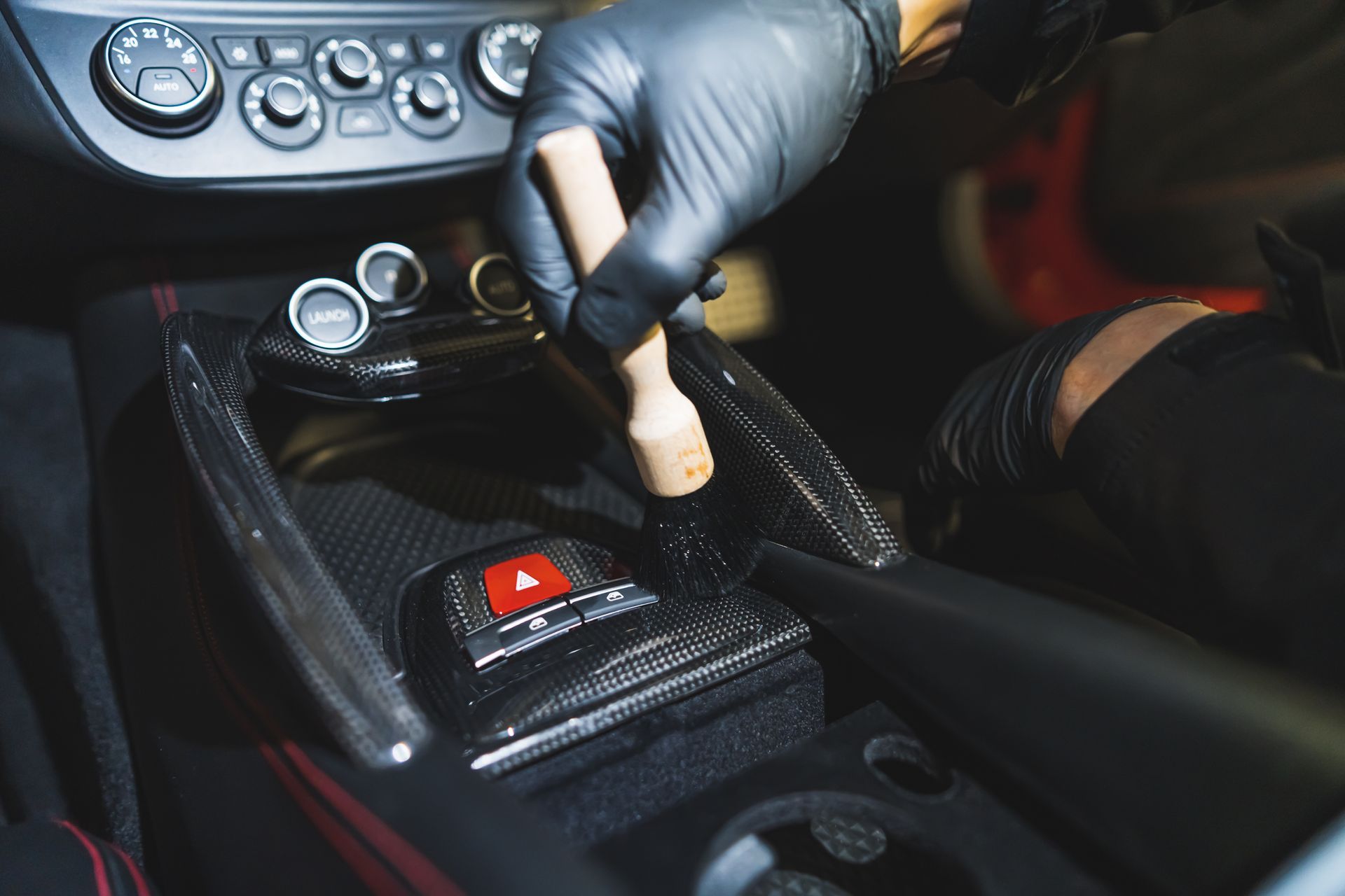 A gloved hand uses a detailing brush to clean a carbon-fiber center console inside a car.