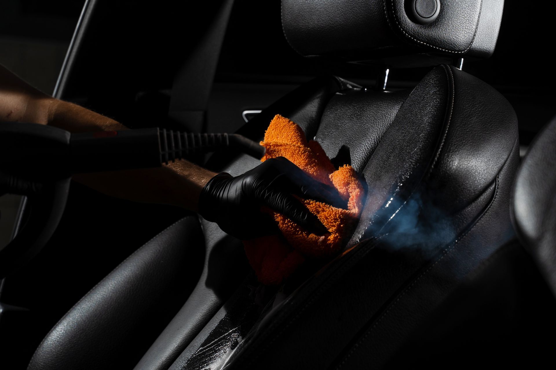 A gloved hand uses a steam nozzle and orange microfiber cloth to deep clean a black leather car seat.