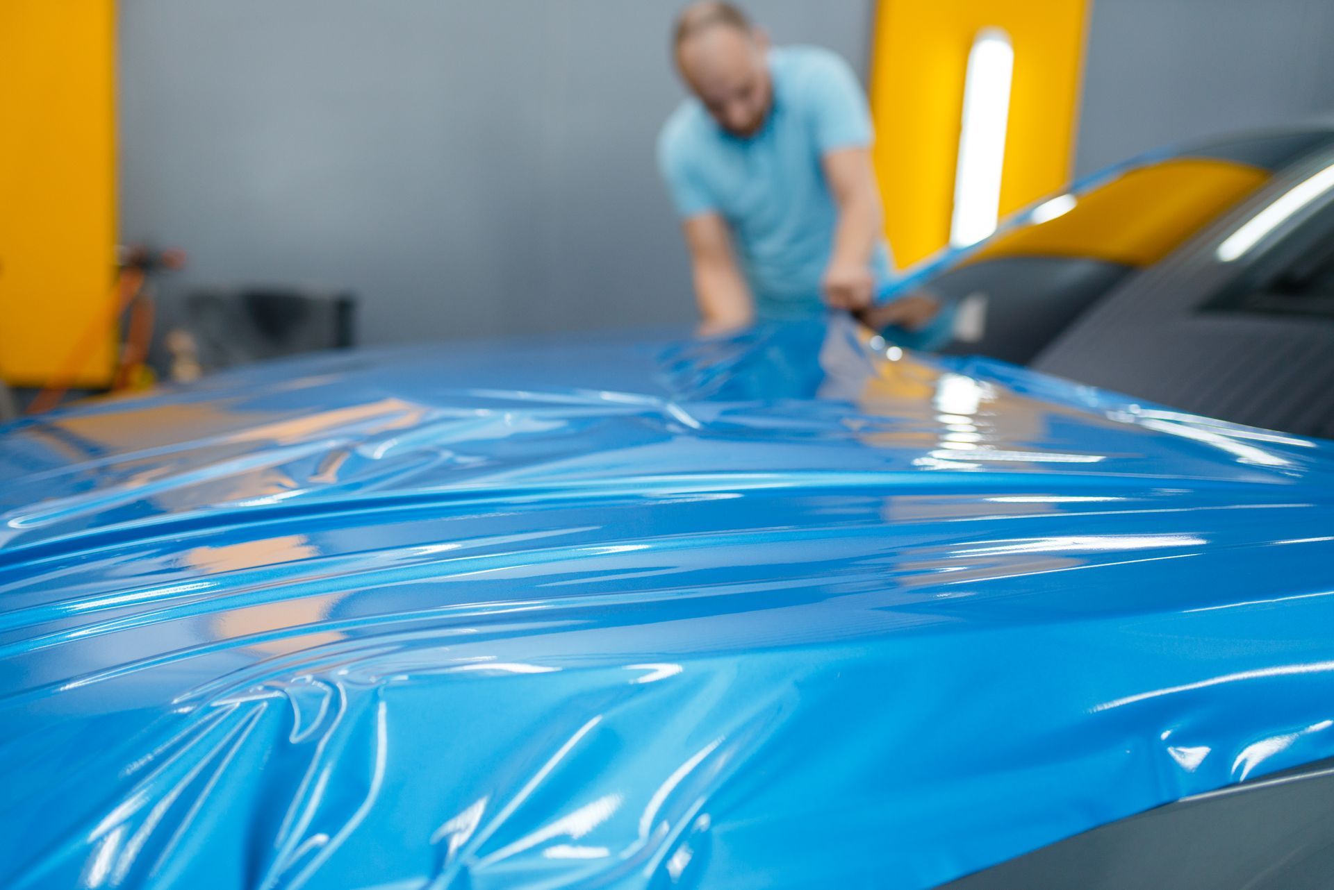A technician applying a glossy blue protective vinyl film to a car hood in a workshop.