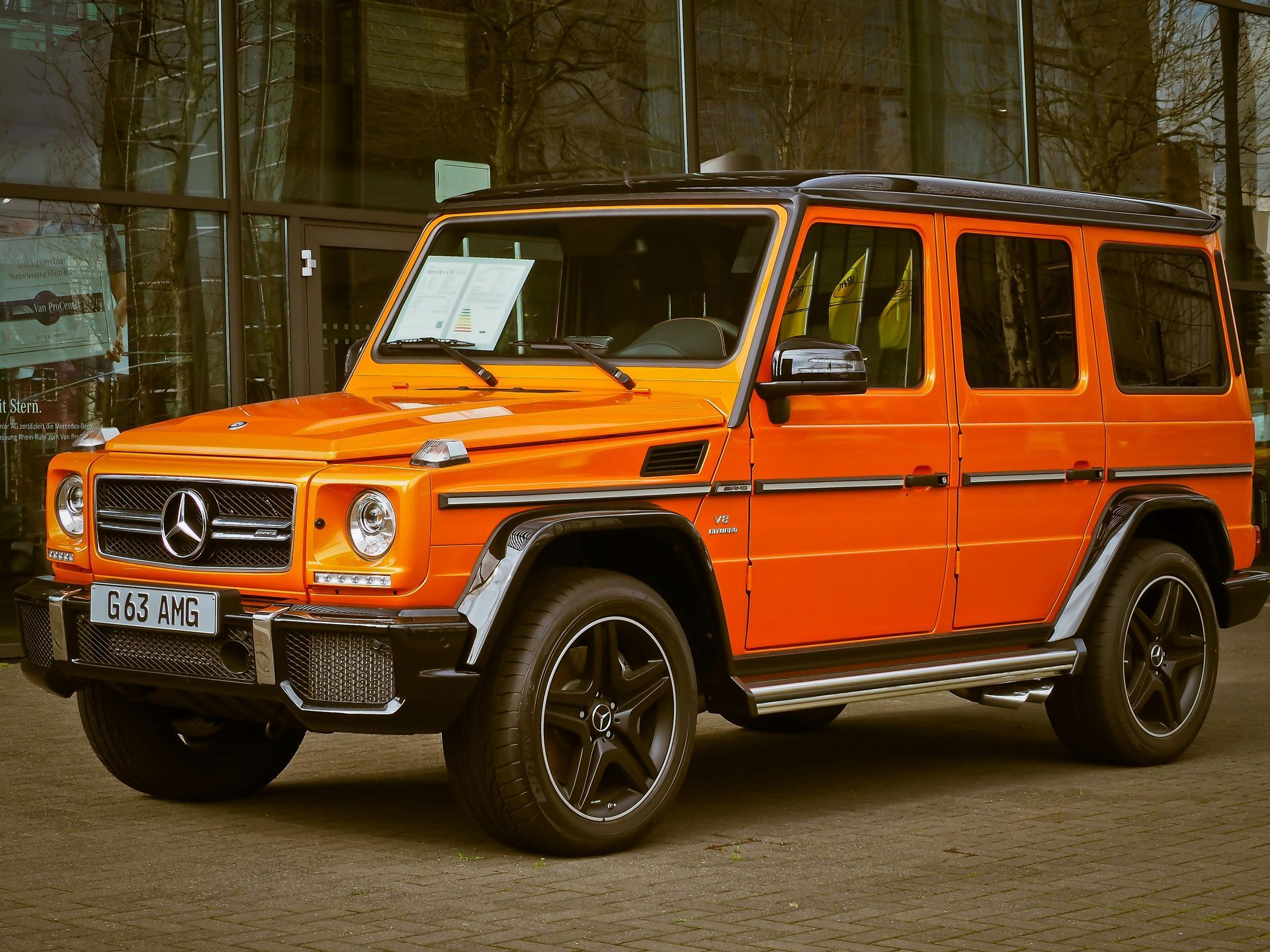 A bright orange Mercedes-Benz G-Wagon parked outdoors in front of a glass-walled building.