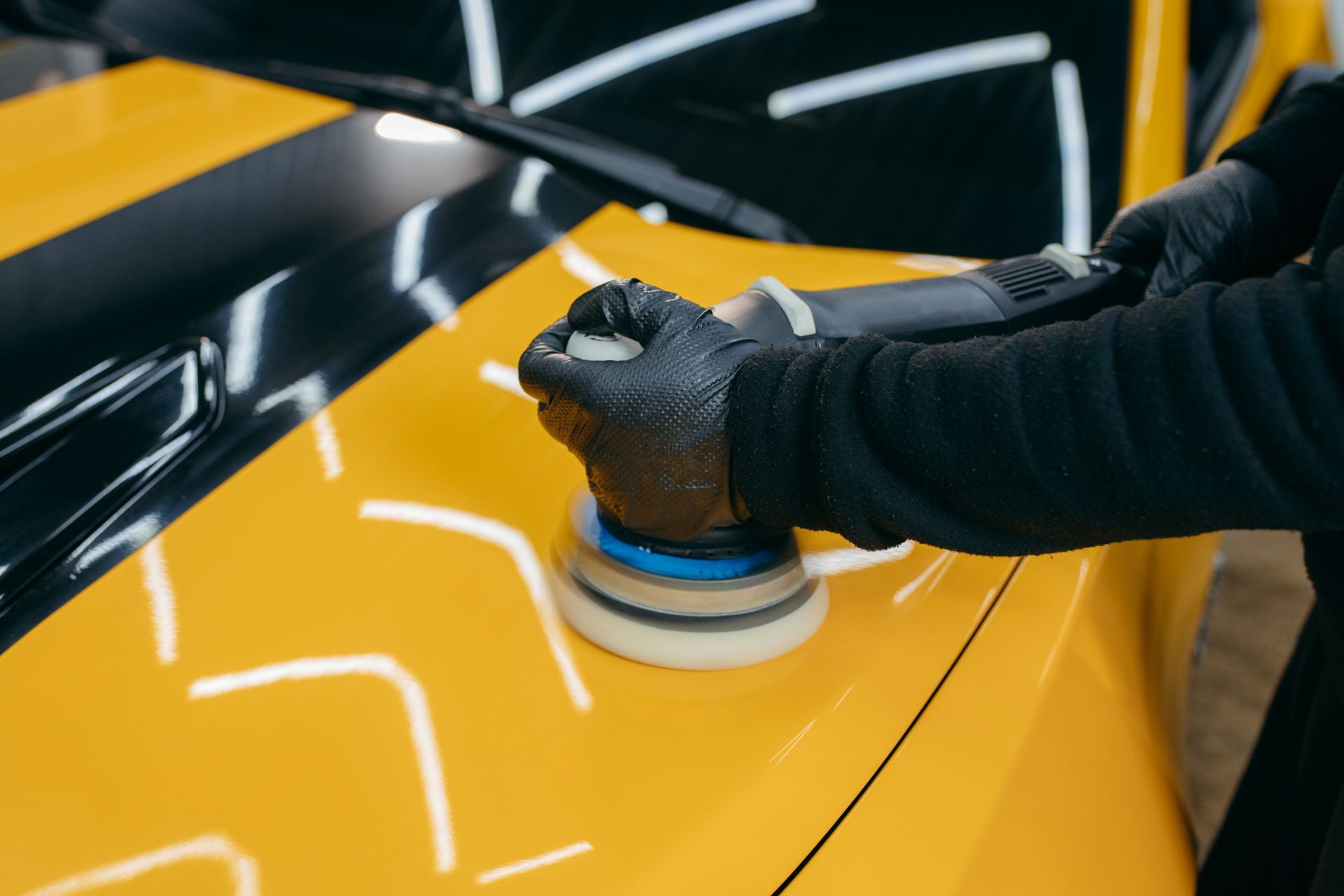 A person wearing black gloves uses a power buffer to polish the bright yellow hood of a vehicle.