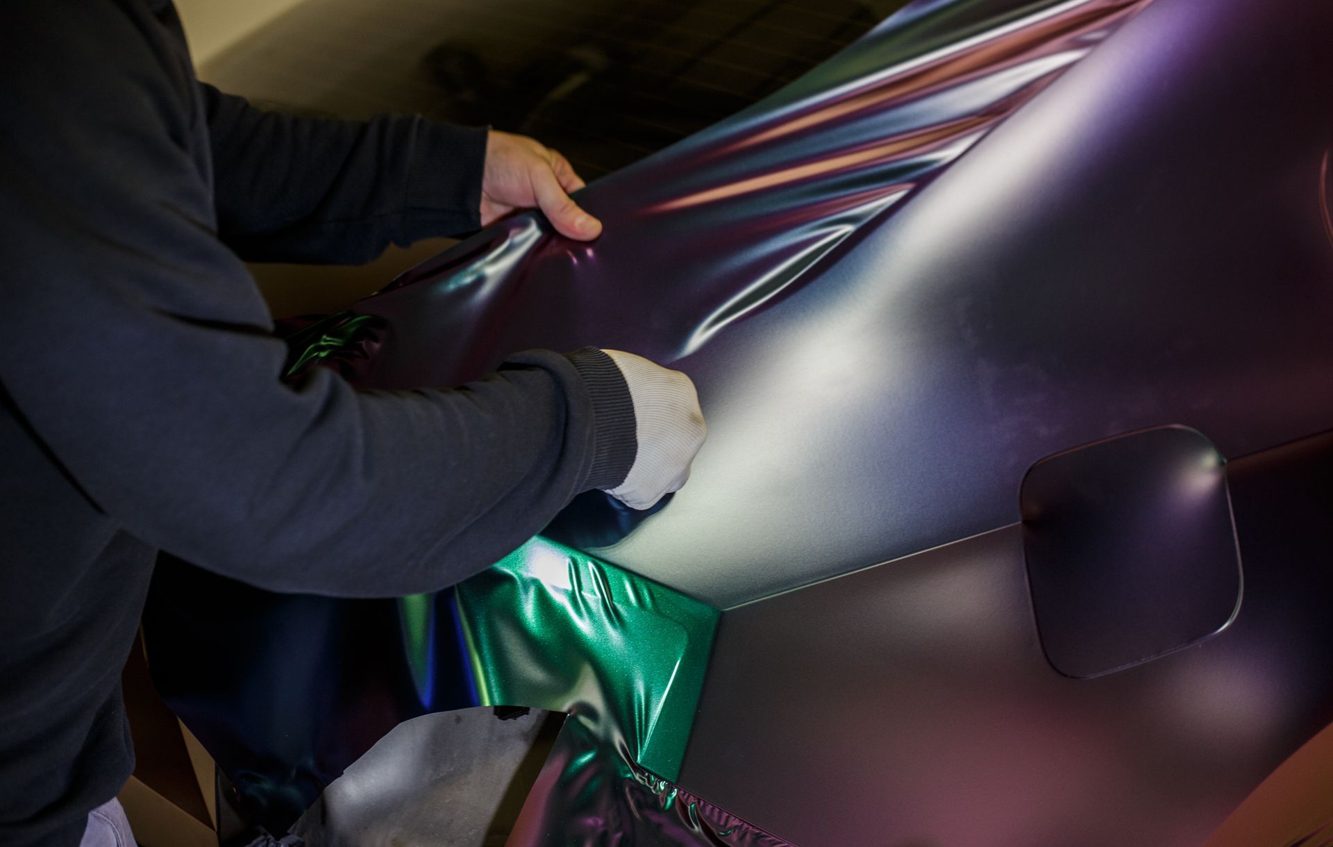 A person applying a color-shifting, iridescent purple and green car wrap to a vehicle's body panel.