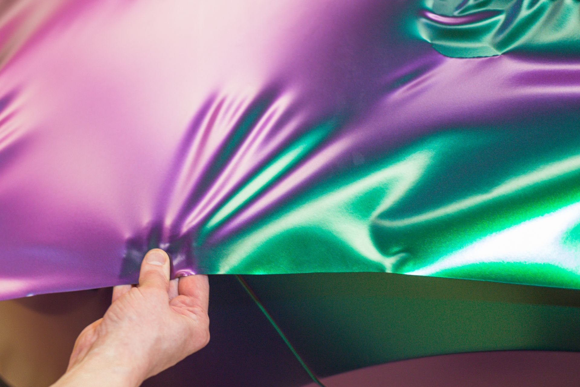 A hand holds iridescent, color-shifting purple and green vinyl wrap over a dark car surface.