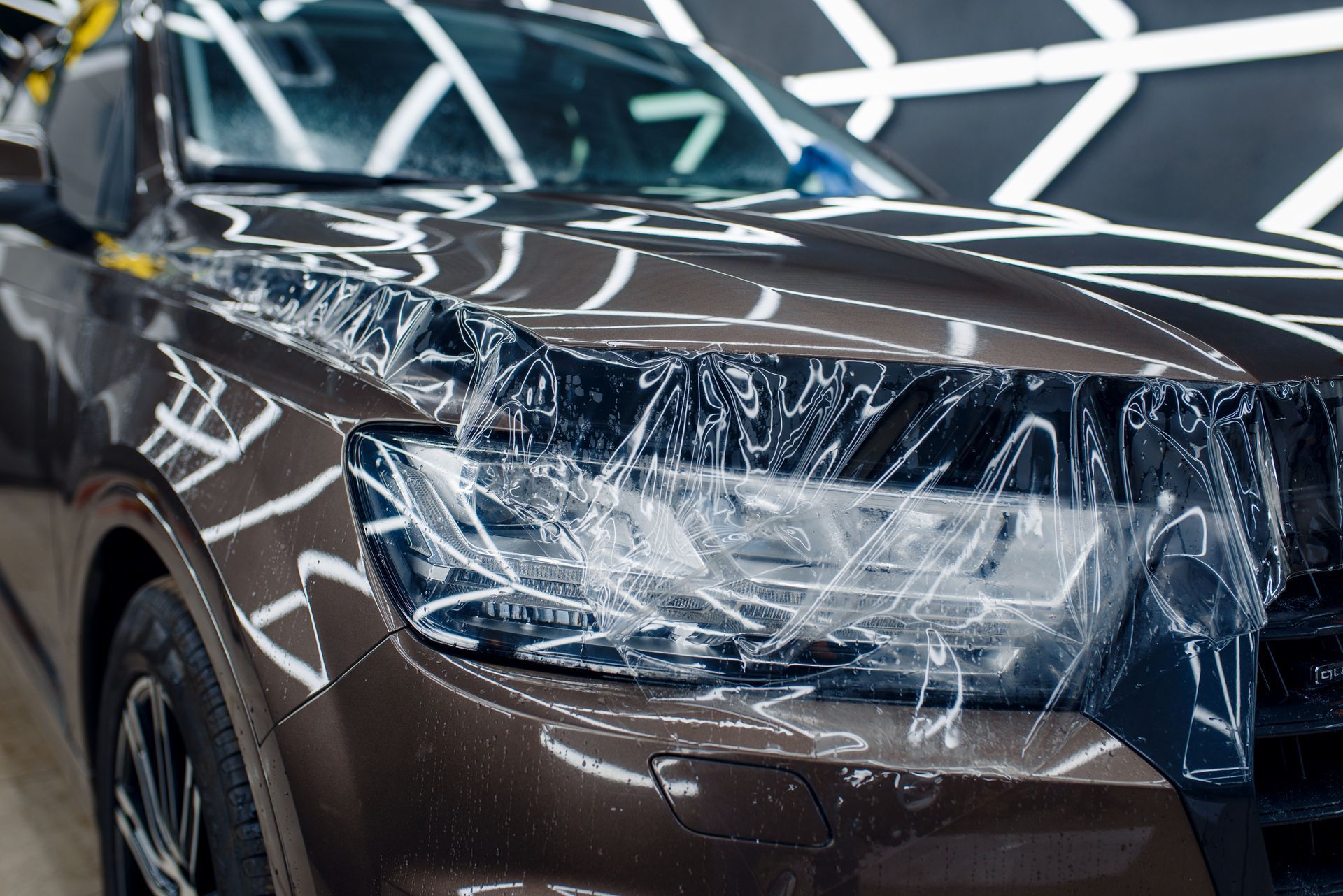 A close-up of a brown car headlight partially covered in protective plastic film in a well-lit workshop.