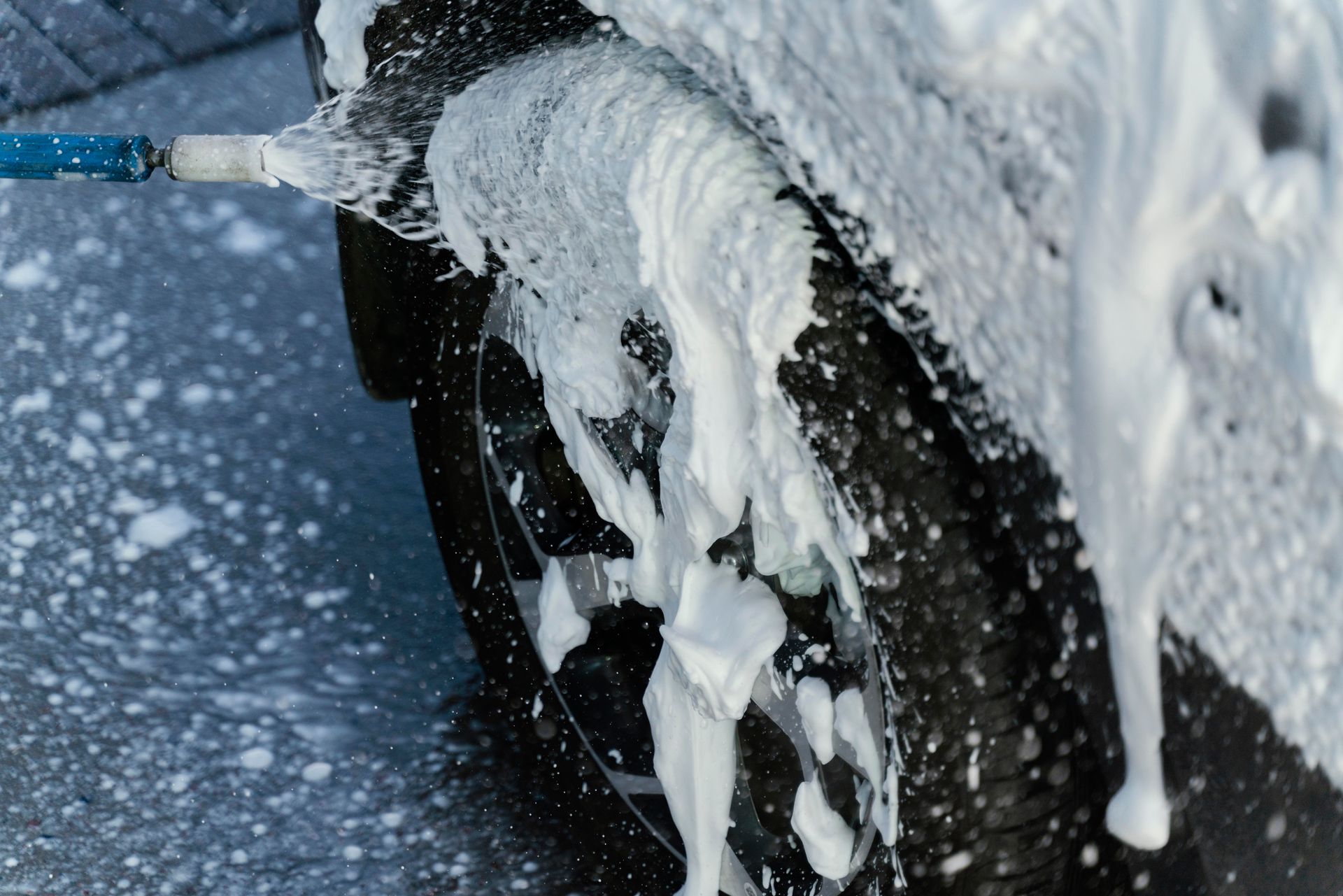 A spray wand sprays thick white soap suds over a car wheel and tire during a wash.