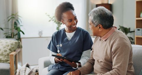 Nurse in blue scrubs reviews tablet with patient in living room.