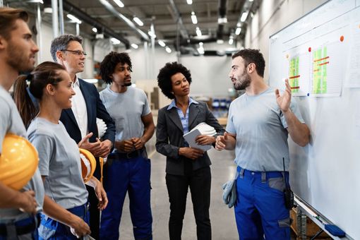A group of people are standing around a whiteboard in a factory.