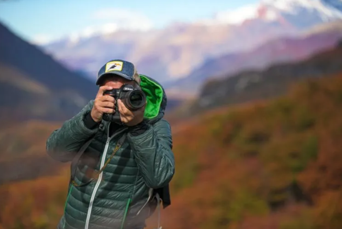 Persona con chaqueta verde tomando una foto con una cámara en un paisaje de montaña.