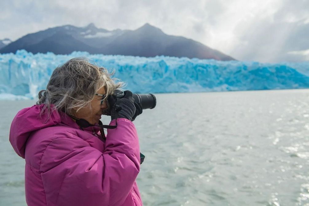 Una persona con una chaqueta magenta fotografía un glaciar con montañas al fondo.