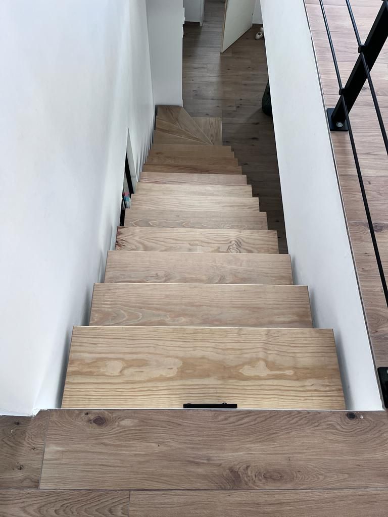 Vue en plongée d'un escalier droit aux marches en bois clair, entre des murs blancs et une rampe en métal noir.