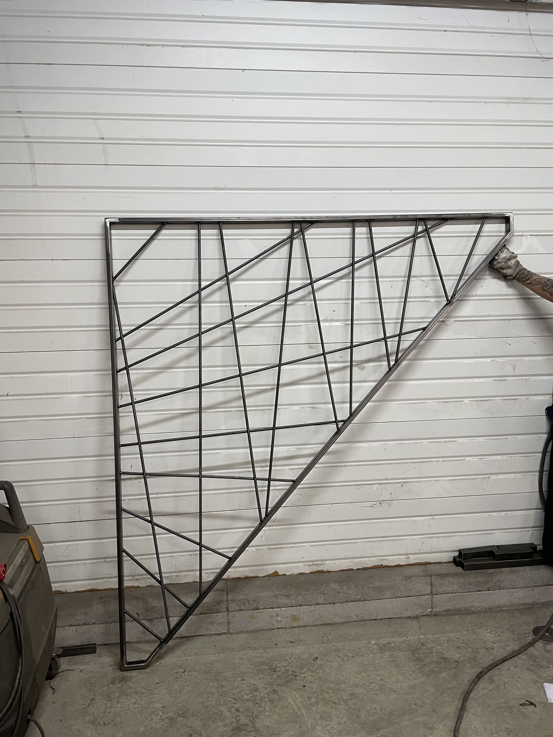 Une personne tenant contre un mur un grand cadre triangulaire en métal soudé sur mesure, orné d'un motif géométrique complexe.