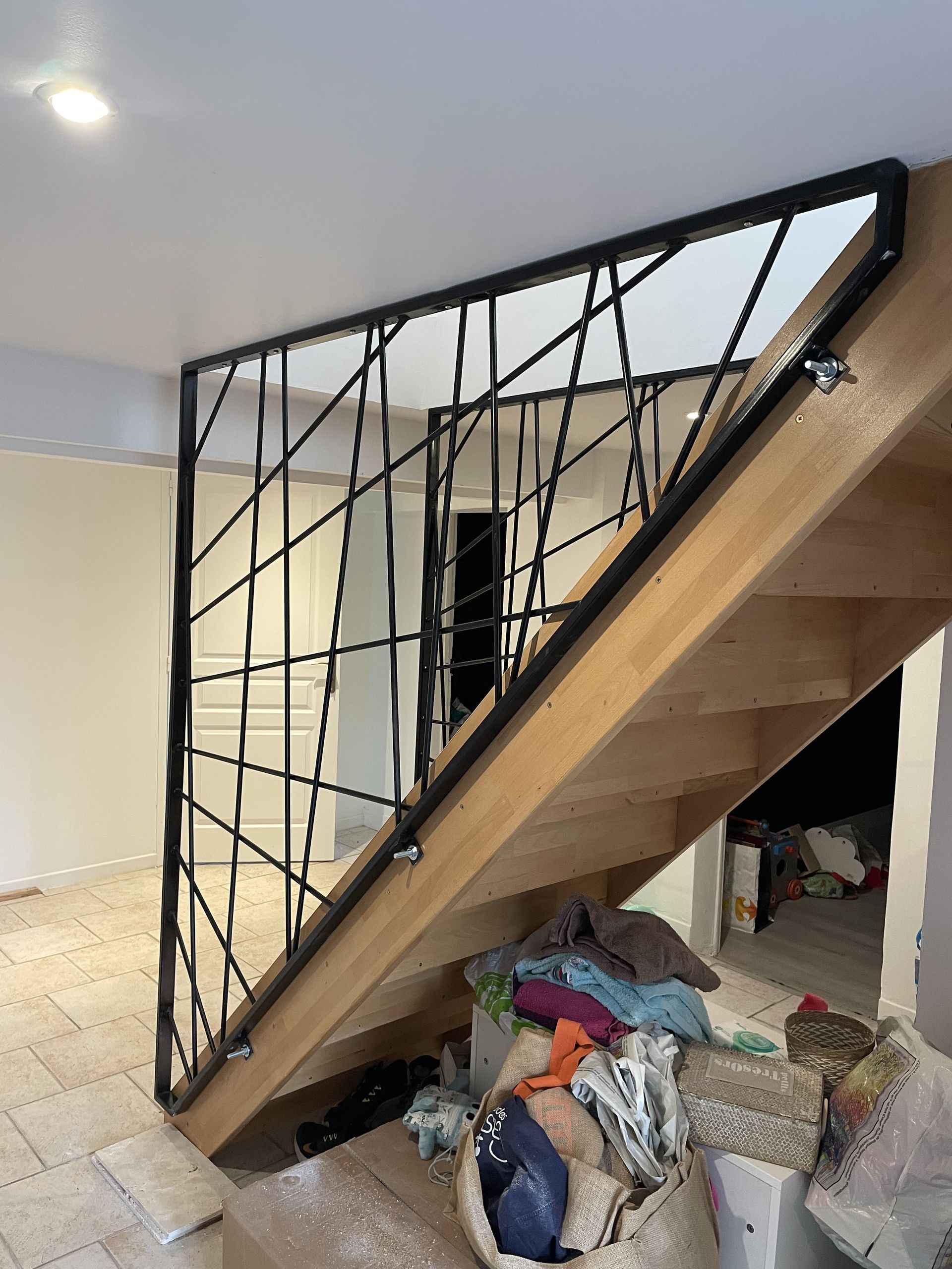 Un escalier en bois avec une rampe moderne en métal noir à géométrie géométrique, placé dans une pièce au sol carrelé.