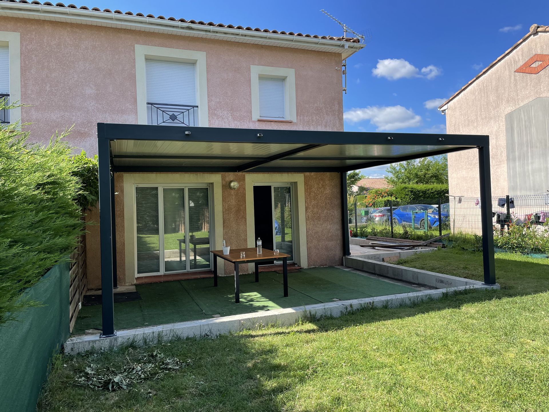 Une pergola en métal noir installée au-dessus d'un patio attenant à une maison en stuc clair.