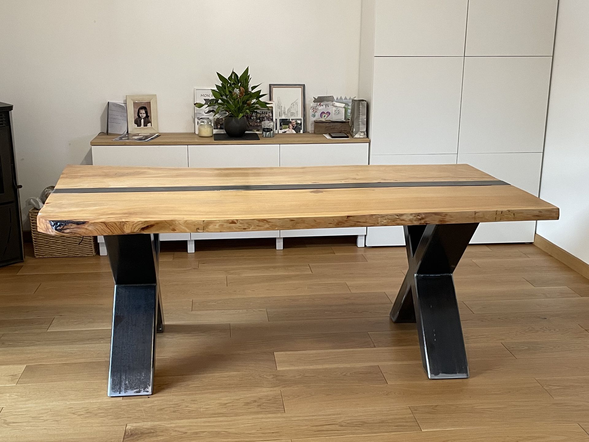 Une table de salle à manger en bois à bords naturels avec une incrustation centrale en résine et des pieds en métal noir en forme de X, posée sur un sol en bois.